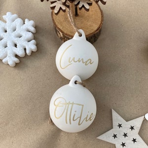 May include: Two white Christmas ornaments with gold lettering, one reading "Luna" and the other "Ottilie". A wooden snowflake Christmas tree and other wooden decorations are also visible.