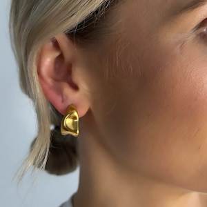 May include: Gold-toned, abstract, irregular shaped hoop earrings. The earrings are worn by a person with blonde hair.