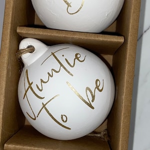 May include: A set of three white, egg-shaped ornaments with gold script. The ornaments are in a brown cardboard box. The text on the ornaments reads "Oh Baby", "Auntie To Be", and "Baby Quinn May 2021". Each ornament has a jute string for hanging.