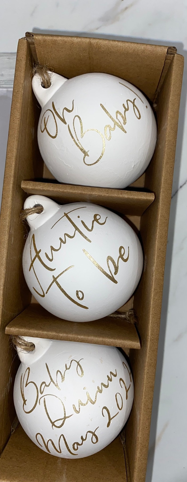 May include: A set of three white, egg-shaped ornaments with gold script. The ornaments are in a brown cardboard box. The text on the ornaments reads "Oh Baby", "Auntie To Be", and "Baby Quinn May 2021". Each ornament has a jute string for hanging.