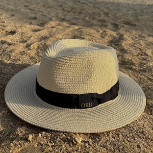 Puede incluir: Sombrero fedora beige con una cinta negra y letras doradas "OKB". El sombrero, de material tejido, tiene un ala ancha. Descansa sobre arena. Un accesorio elegante para días soleados.