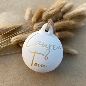 May include: A white ceramic ornament with gold lettering that reads "Lauren & Tom". The ornament is round with a small loop for hanging and is suspended by a natural twine. Dried, tan-colored grasses are in the background.