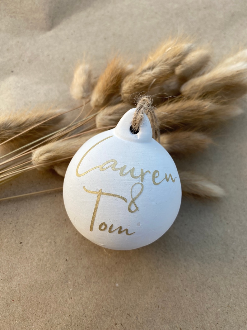 May include: A white ceramic ornament with gold lettering that reads "Lauren & Tom". The ornament is round with a small loop for hanging and is suspended by a natural twine. Dried, tan-colored grasses are in the background.