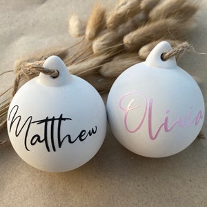 May include: Two white ceramic Christmas ornaments with names Matthew and Olivia in black and pink script. Each ornament has a jute twine loop for hanging. Dried bunny tail grass is in the background.
