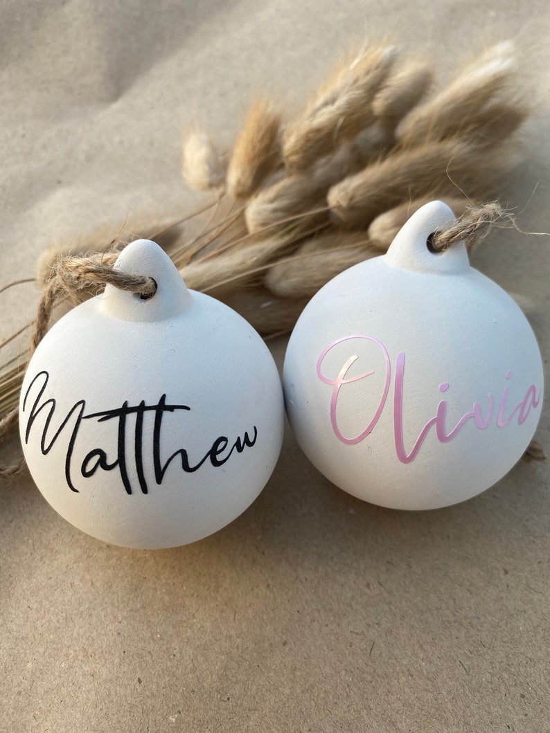 May include: Two white ceramic Christmas ornaments with names Matthew and Olivia in black and pink script. Each ornament has a jute twine loop for hanging. Dried bunny tail grass is in the background.