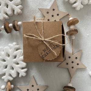 May include: A small, square, brown gift box tied with twine and a tag that reads "MERRY CHRISTMAS." The box is surrounded by wooden star ornaments with black star cutouts, white snowflakes, and a garland of wooden beads and slices.