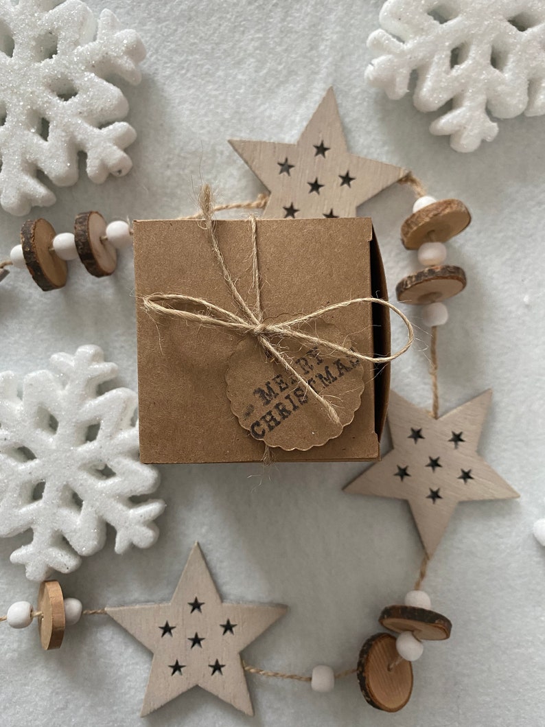 May include: A small, square, brown gift box tied with twine and a tag that reads "MERRY CHRISTMAS." The box is surrounded by wooden star ornaments with black star cutouts, white snowflakes, and a garland of wooden beads and slices.