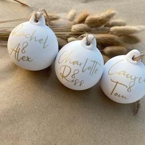 May include: Three white ceramic ornaments with gold lettering and twine hangers. The ornaments are personalized with names: Rachel & Alex, Charlotte Ross, and Lauren & Tom. The background includes dried flowers.