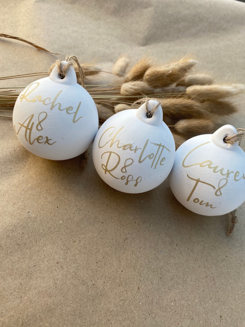 May include: Three white ceramic ornaments with gold lettering and twine hangers. The ornaments are personalized with names: Rachel & Alex, Charlotte Ross, and Lauren & Tom. The background includes dried flowers.