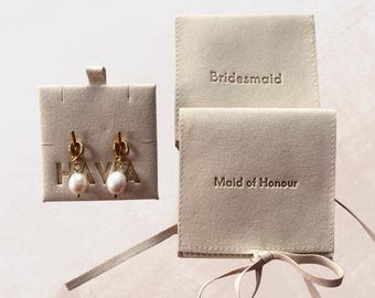 May include: A pair of gold-toned earrings with white pearl drops attached to a beige display card labeled HAVA, alongside two beige velvet pouches with the text Bridesmaid and Maid of Honour embossed on the front flaps with ribbon ties.