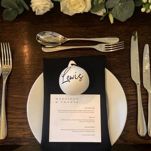 May include: A formal table setting with a white plate, black napkin, and silverware. A menu card reads "Maddison & Thomas" with a list of courses. A white ornament with the name "Lewis" is on top of the napkin. White flowers and greenery are in the background.