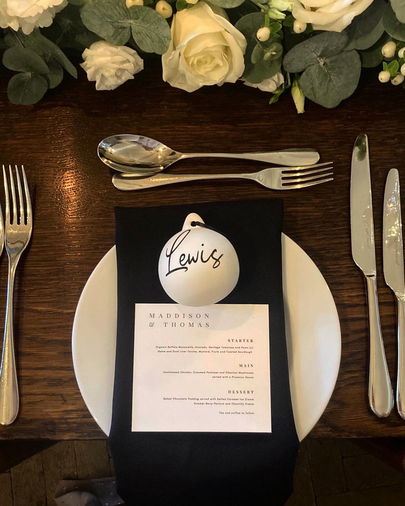 May include: A formal table setting with a white plate, black napkin, and silverware. A menu card reads "Maddison & Thomas" with a list of courses. A white ornament with the name "Lewis" is on top of the napkin. White flowers and greenery are in the background.