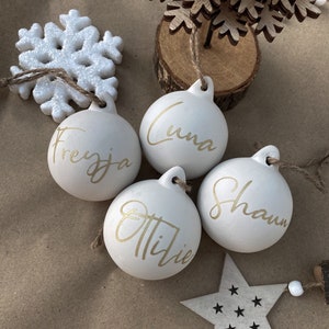 May include: Four white Christmas ornaments with gold lettering spelling out names: Freyya, Luna, Ottilie, and Shaun. A white glitter snowflake, a wooden star, and a small wooden tree are also visible.