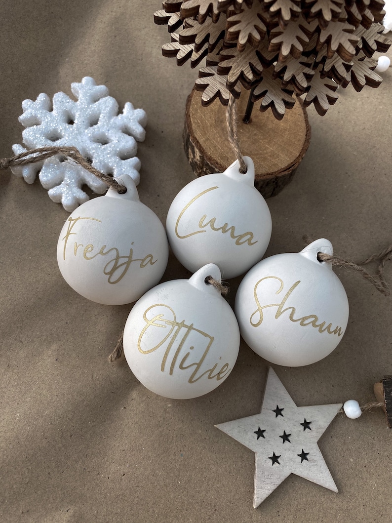 May include: Four white Christmas ornaments with gold lettering spelling out names: Freyya, Luna, Ottilie, and Shaun. A white glitter snowflake, a wooden star, and a small wooden tree are also visible.