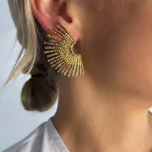 Peut inclure: Boucles d'oreilles statement dorées à l'aspect éventail. Les boucles d'oreilles sont en métal fin texturé.