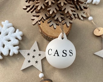 May include: A white Christmas ornament with the name "CASS" in black lettering. The ornament is surrounded by wooden star and snowflake decorations, a small wooden Christmas tree, and a garland of wooden beads and discs. The background is a light brown surface.
