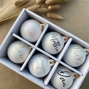 May include: A white gift box containing six personalized Christmas ornaments. Each ornament has a name in a different color: pink, black, gold, and silver. The ornaments are round with a small loop for hanging.