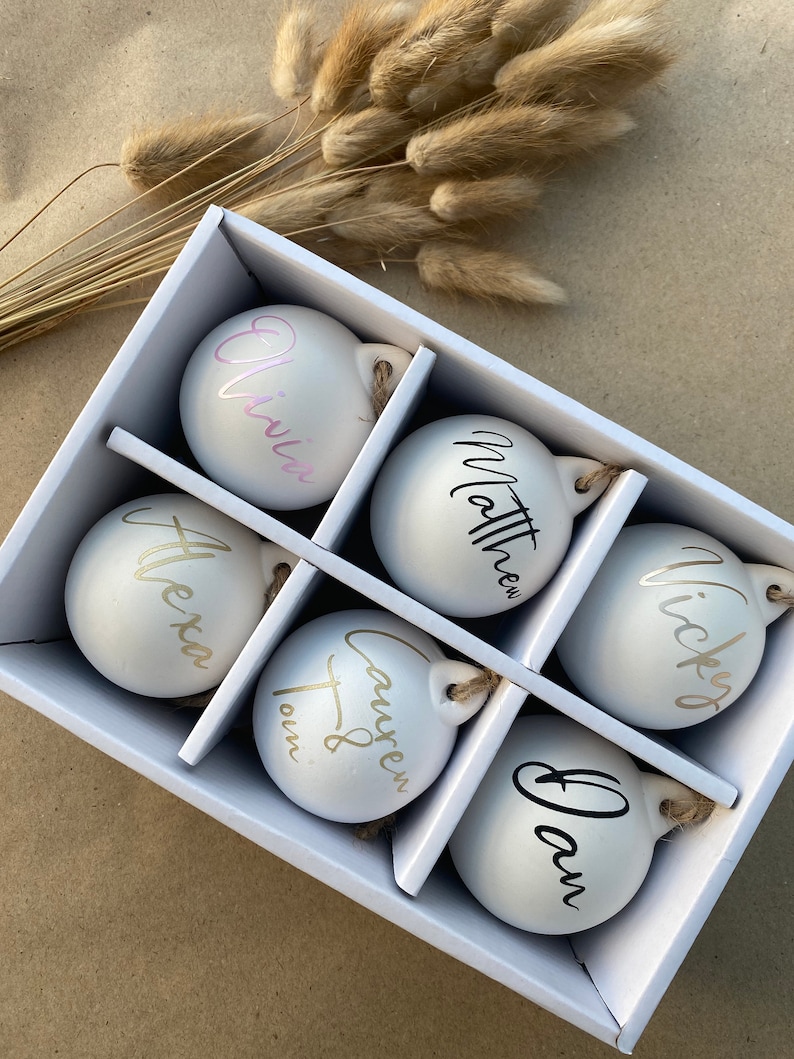 May include: A white gift box containing six personalized Christmas ornaments. Each ornament has a name in a different color: pink, black, gold, and silver. The ornaments are round with a small loop for hanging.