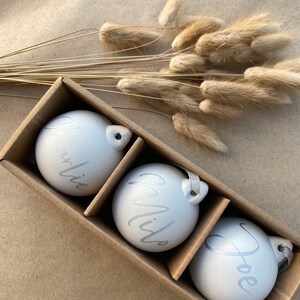 May include: Three white Christmas ornaments with silver lettering spelling out "Charlie", "Milo", and "Joe" are displayed in a cardboard box. Each ornament has a silver ribbon for hanging. Dried, tan-colored bunny tail grass is in the background.