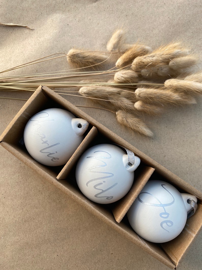 May include: Three white Christmas ornaments with silver lettering spelling out "Charlie", "Milo", and "Joe" are displayed in a cardboard box. Each ornament has a silver ribbon for hanging. Dried, tan-colored bunny tail grass is in the background.