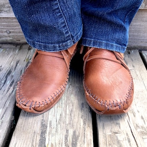 Peut inclure: Une paire de mocassins en cuir marron avec des coutures d&eacute;coratives. Les mocassins sont port&eacute;s par une personne portant un jean bleu.