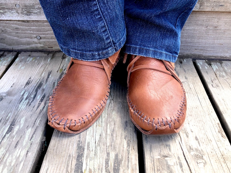Peut inclure: Une paire de mocassins en cuir marron avec des coutures d&eacute;coratives. Les mocassins sont port&eacute;s par une personne portant un jean bleu.