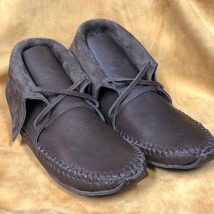May include: A pair of brown leather moccasin-style shoes. The shoes have a rounded toe, stitched seams, and a suede collar. Leather laces are tied across the top. The shoes are displayed on a golden-yellow surface.