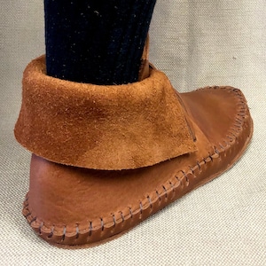 Peut inclure: Gros plan d'une mocassine en cuir marron avec un revers en daim. La mocassine est cousue avec une couture d&eacute;corative autour de la semelle.