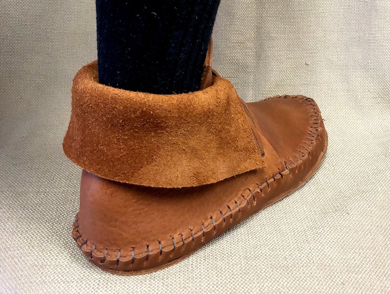 Peut inclure: Gros plan d'une mocassine en cuir marron avec un revers en daim. La mocassine est cousue avec une couture d&eacute;corative autour de la semelle.