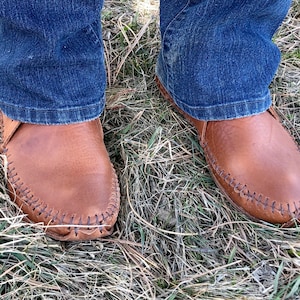 Peut inclure: Une paire de mocassins en cuir marron avec une couture sur le bord. Les mocassins sont port&eacute;s par une personne portant un jean bleu.