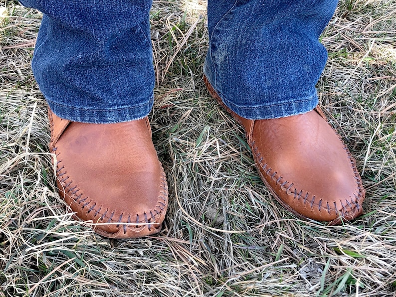 Peut inclure: Une paire de mocassins en cuir marron avec une couture sur le bord. Les mocassins sont port&eacute;s par une personne portant un jean bleu.