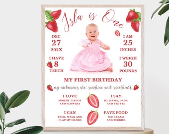 Op de afbeelding: Een roze en witte eerste verjaardag mijlpaalposter met een foto van een babymeisje in een roze jurk. De poster heeft een aardbeien thema met rode aardbeien en groene bladeren. De poster bevat de naam van de baby, Isla, en details over haar eerste jaar, inclusief haar geboortedatum, lengte, gewicht en favoriete dingen.