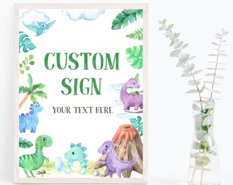 Op de afbeelding: Een printbaar bord met een witte achtergrond en een groene rand. Het bord heeft een cartoon-dinosaurusthema met een vulkaan, palmbomen en dinosaurussen in verschillende kleuren. De tekst "CUSTOM SIGN" staat in groene letters en de tekst "YOUR TEXT HERE" staat in grijze letters.