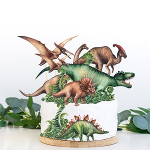 Könnte beinhalten: Ein weißer Kuchen, der mit grünen Farnen und fünf verschiedenen Dinosaurierfiguren verziert ist. Die Dinosaurier sind ein grüner T-Rex, ein brauner Stegosaurus, ein brauner Triceratops, ein grüner Pterodactylus und ein brauner Brachiosaurus.