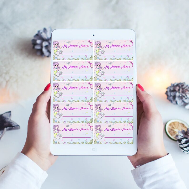 May include: A sheet of printable labels featuring a cartoon mermaid with pink hair and a purple tail. The labels are pink with a green and blue floral border. The text on the labels reads "My Mermaid Name is".