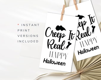 May include: Two white Halloween-themed prints with the phrases "Creep It Real" and "Creep It Real Happy Halloween" in black script. The prints are held by wooden clips, with a white background and a hint of dried palm leaves.