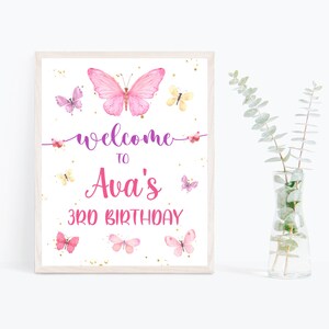 May include: A white sign with a pink butterfly and gold glitter. The sign says "Welcome to Ava's 3rd Birthday".