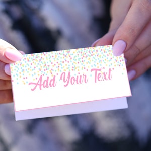 Op de afbeelding: Een kleine, gevouwen witte kaart met de tekst "Add Your Text" in roze letters. De bovenrand is versierd met een confetti-patroon in pastelkleuren. De kaart wordt tussen twee handen gehouden.