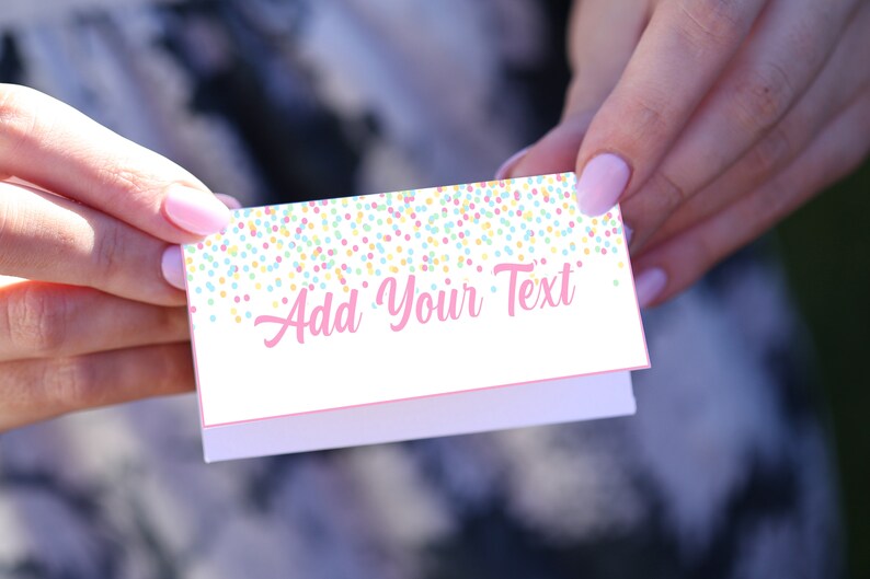 Op de afbeelding: Een kleine, gevouwen witte kaart met de tekst "Add Your Text" in roze letters. De bovenrand is versierd met een confetti-patroon in pastelkleuren. De kaart wordt tussen twee handen gehouden.