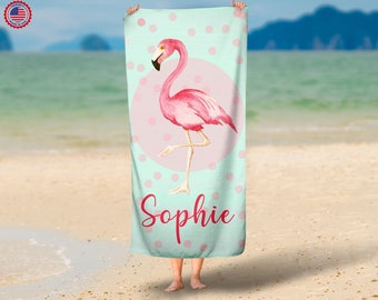May include: A beach towel featuring a pink flamingo illustration on a light blue background with pink polka dots. The flamingo stands on one leg within a pink circle. The name "Sophie" is written in red script below the flamingo.