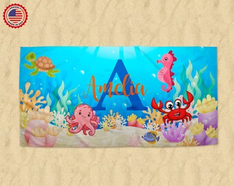 May include: A personalized beach towel featuring an underwater scene with cartoon sea creatures. The design includes a turtle, seahorse, octopus, crab, and fish, with the name "Amelia" and a large letter "A" in the center. The towel is made in the USA.