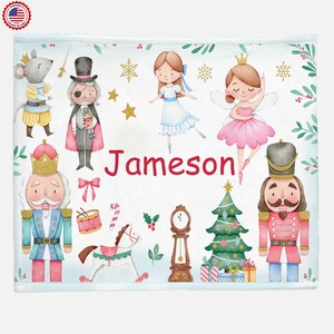 May include: White blanket featuring a watercolor illustration of characters from The Nutcracker ballet, including a mouse, a nutcracker, and ballerinas. The name "Jameson" is written in red. The blanket is decorated with Christmas-themed elements.
