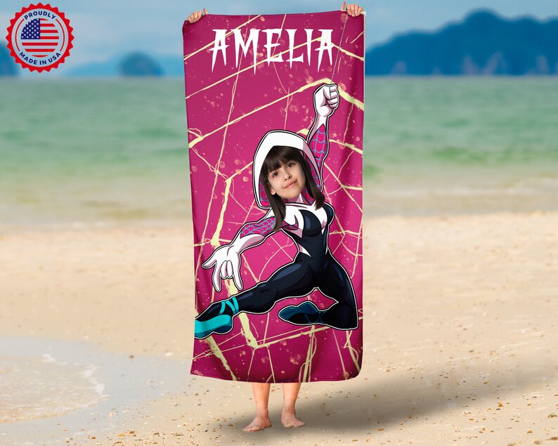 May include: A pink beach towel with a spider-woman design. The design features a woman in a white and black suit with a pink spider symbol on her chest. The towel has the name "Amelia" printed at the top.