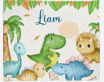 May include: A white baby blanket with a green, blue, and yellow dinosaur design. The blanket has the name "Liam" written in blue cursive letters at the top.