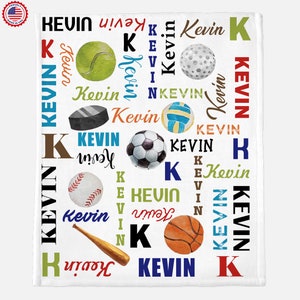 May include: White baby blanket with a colourful pattern of sports balls and the name Kevin repeated throughout. The sports balls include a baseball, basketball, football, tennis ball, golf ball, hockey puck, and a volleyball.