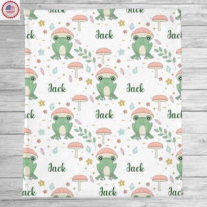 May include: A white baby blanket with a repeating pattern of green frogs sitting on pink mushrooms with white dots. The background is filled with small flowers, crystals, and leaves. The name "Jack" is repeated throughout the pattern.