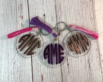 May include: Three clear acrylic keychains with silver metal rings and colorful tassels. Each keychain features a different monogram: "AME" in purple, "AME" in silver, and "CMM" in silver.