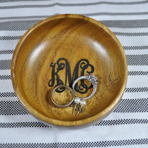Może przedstawiać: Drewniana miska z wygrawerowanym czarnym monogramem "KMC" wewnątrz. Miska jest wypełniona biżuterią, w tym pierścionkiem, kolczykami i bransoletką.