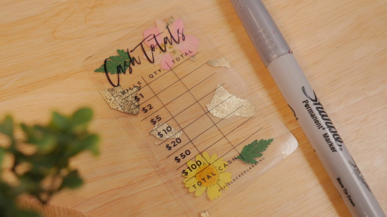 May include: A clear plastic cash register card with a floral design. The card has a grid for counting bills and a space for the total cash amount. The text "Cash Totals" is written in black cursive at the top of the card.