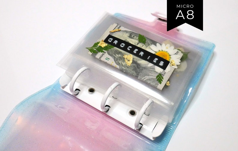 以下が含まれることがあります： 虹色のグリッターカバーが付いた透明なプラスチック製のA8マイクロバインダー。バインダーには、ドル紙幣、ドライフラワー、そして「GROCERIES」と書かれたラベルが入った透明なポケットがあります。